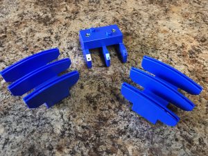 Blue plastic construction piece with four leg-like prongs on a speckled countertop, surrounded by curved blue track sections (toy parts).