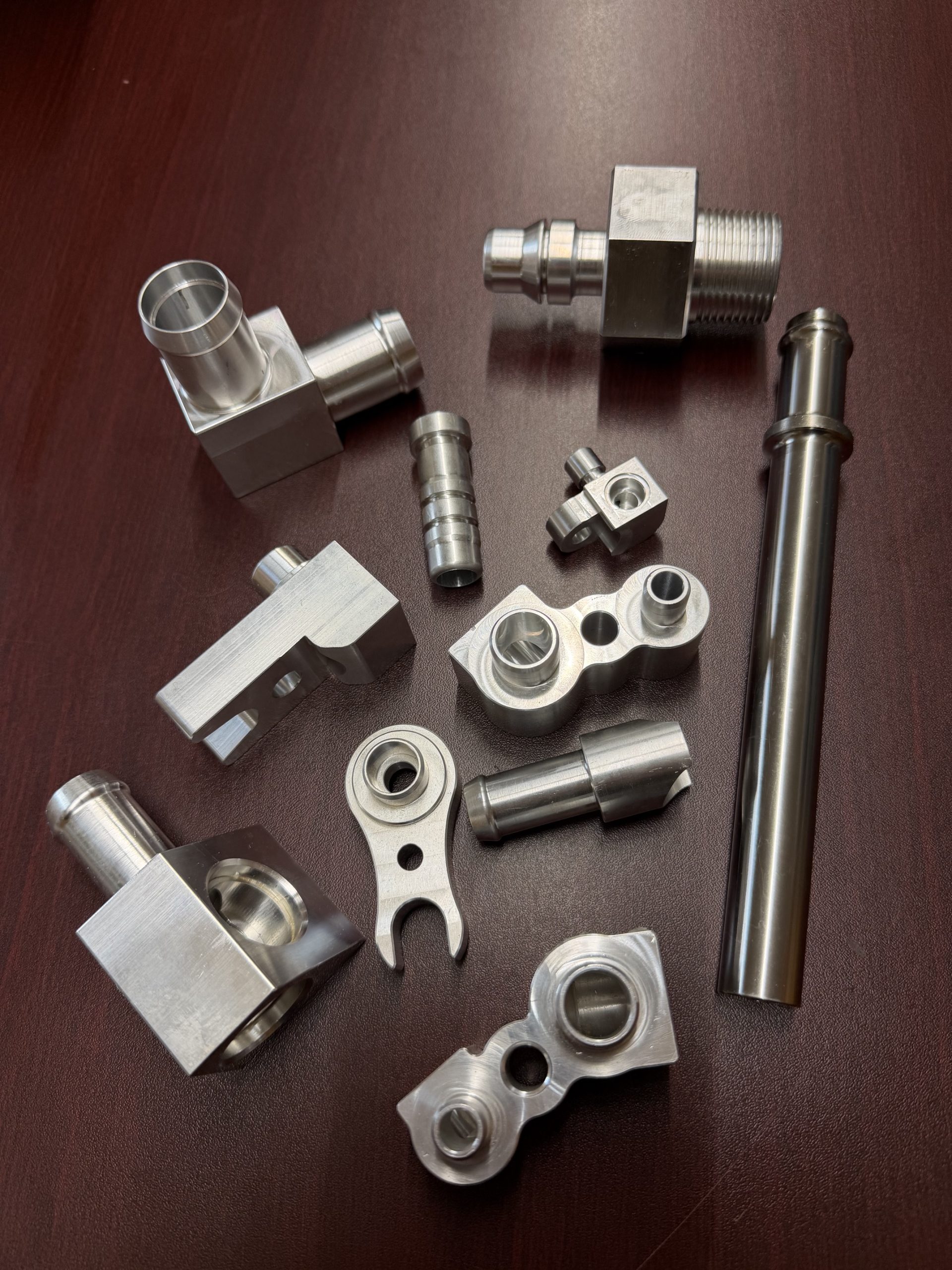 Assorted silver metal fittings and brackets laid out on a dark brown surface, including threaded connectors and a long cylindrical rod.