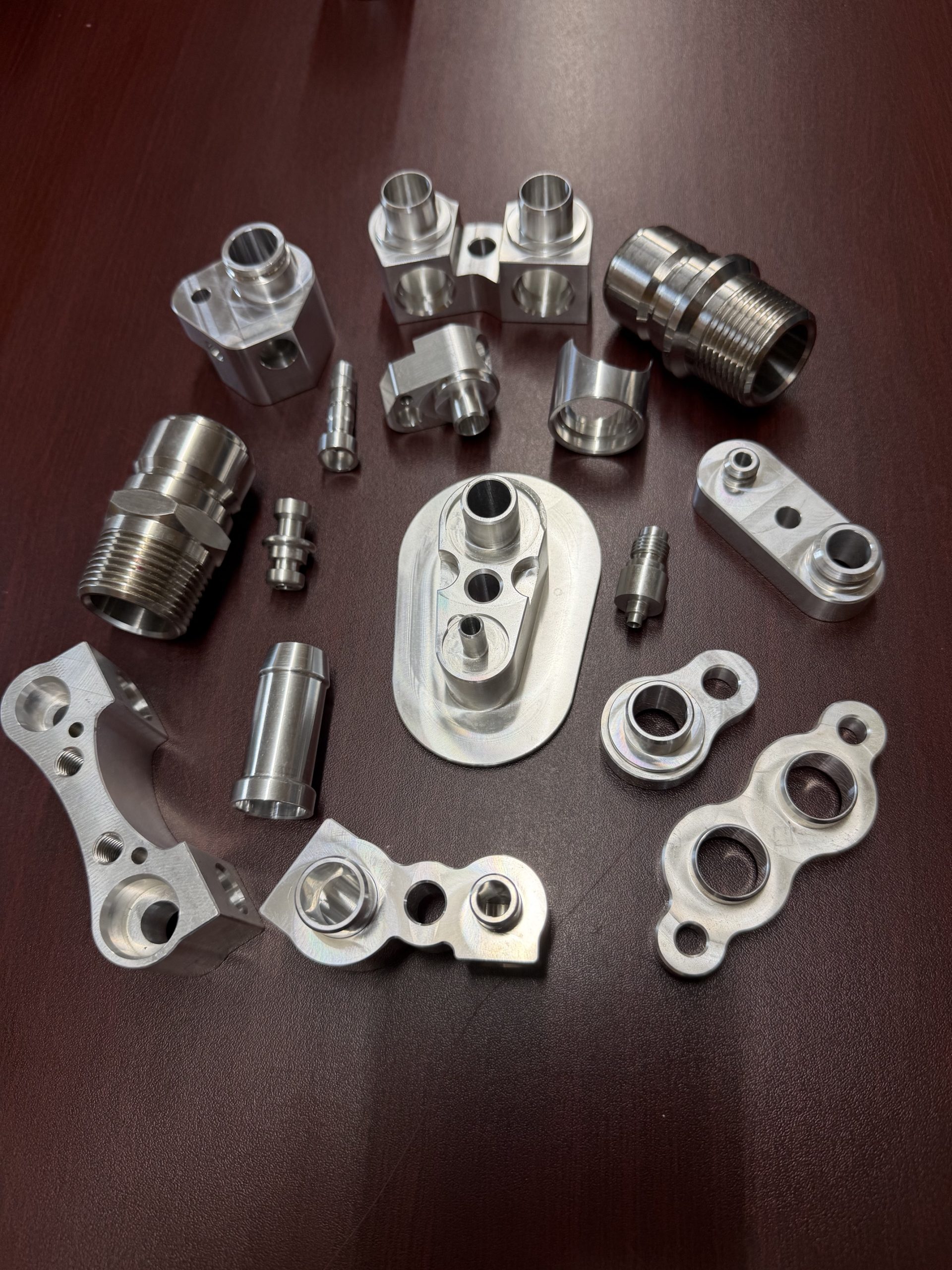 Assorted machined metal fittings and brackets laid out on a dark brown surface.