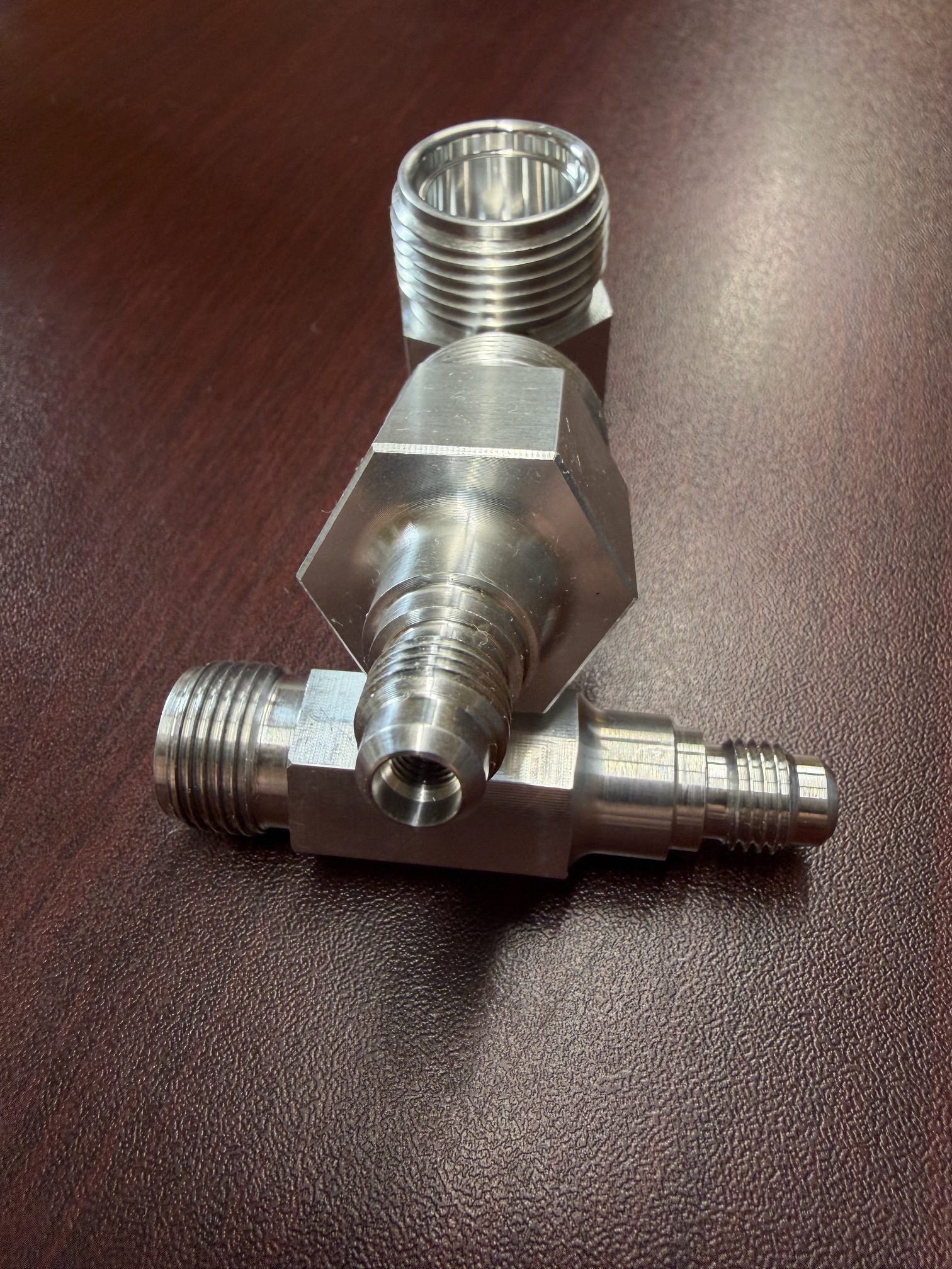 Metal machined adapter fitting with multiple threaded ends, positioned on a brown textured surface